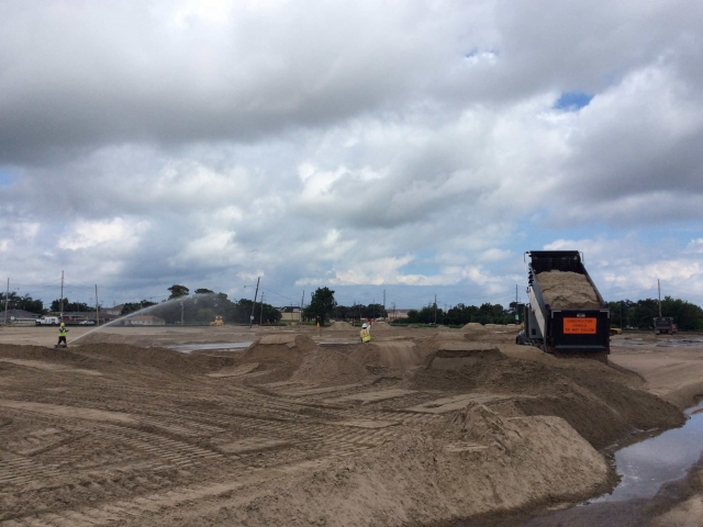 a worker sprays water on the sandy jobsite as another worker guides a dump truck full of sand
