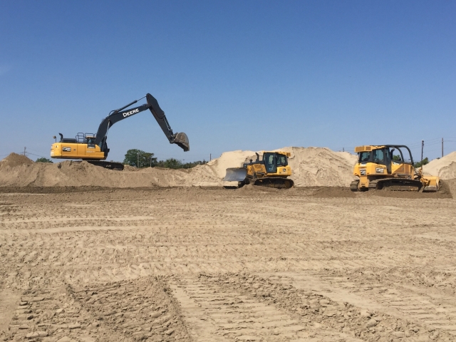 two dozers push sand across the job site while an excavator in the background swings its arm