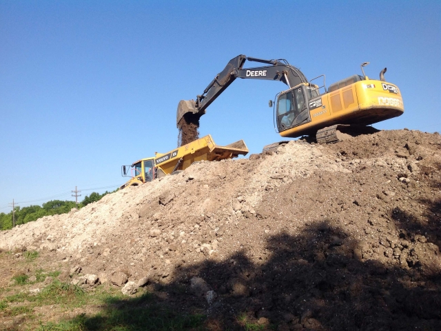 an excavator, on top of a hill, loads a large truck with dirt